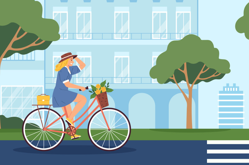Kobieta w kapeluszu i niebieskiej sukience jedzie na rowerze miejską ulicą. W koszyku wiezie kwiaty.